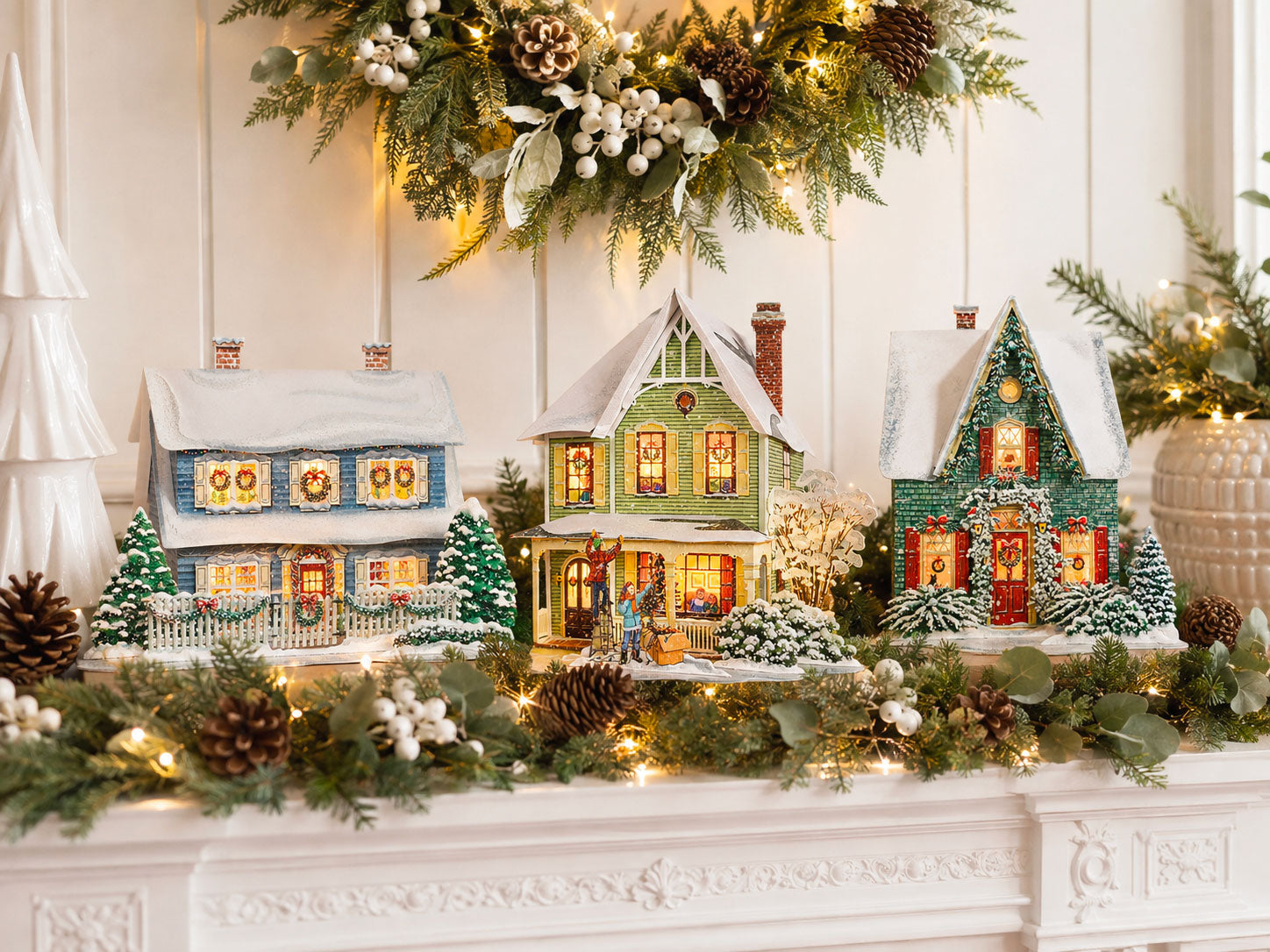 Three CranberryLand Christmas village houses illustrated by Liette Gariépy, displayed on a bright holiday mantel with greenery and warm lights.